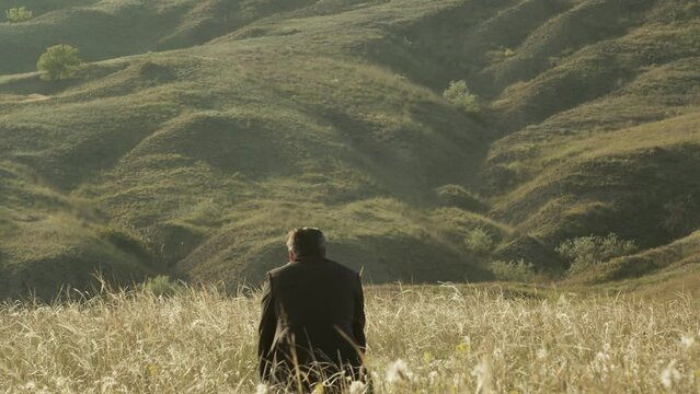 A tired and lonely adult man in a suit in dry grass in front of a green foothill sits with his head down. Back view