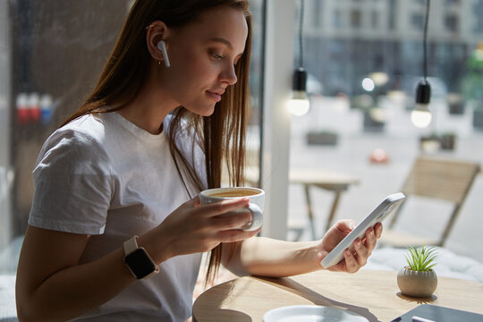 Beautiful young woman drinking coffee, listening to music and using a mobile app on a smart phone in a cafe. Portrait of a cheerful white female in wireless headphones browsing an application