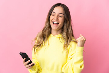Young Romanian woman isolated on pink background using mobile phone and doing victory gesture