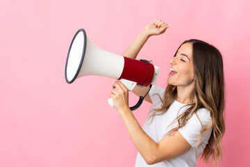 Young Romanian woman isolated on pink background shouting through a megaphone to announce something in lateral position