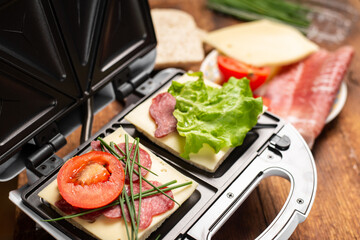 A sandwich maker with two different sandwiches with tomatoes, ham, onions and cheese. Preparing a light meal.