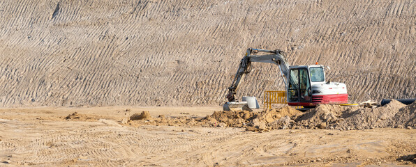 Small white and red excavator moving earth in the middle of a large construction site to build a new road and access to the city. Urban reconstruction works. © sirbouman