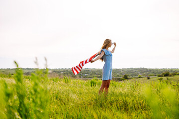 Young woman waving American flag on blooming meadow. 4th of July. Independence Day. Patriotic holiday. USA flag fluttering in the wind.