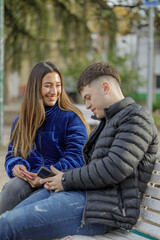 Couple of girl and boy sitting on the bench in a public park look at the mobile phone.