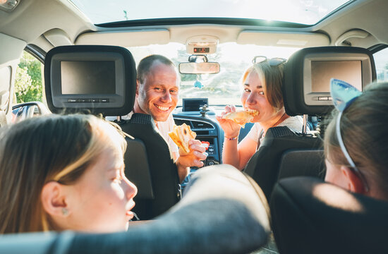 Happy smiling young couple with two daughters eating just cooked Italian pizza sitting in modern car. Happy family moments, childhood, fast food eating or auto journey lunch break concept image..