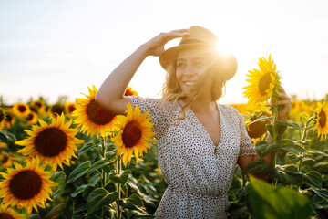 Beautiful woman posing in a field of sunflowers in a dress and hat.  Fashion, lifestyle, travel and vacations concept. - obrazy, fototapety, plakaty