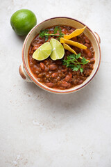 Bowl of chili con carne or spicy meat stew served with lime wedges and nachos, vertical shot on a light-beige stone background, copy space