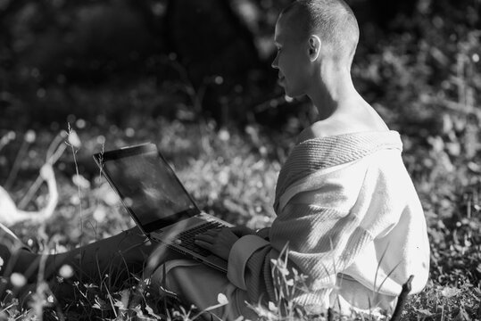 A bald woman in a bathrobe sits on green grass with a laptop in her hands. Online work, freelance concept. Sunny day.Black and white image.