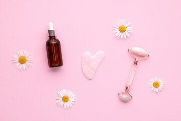 Obraz na płótnie Canvas Quartz face roller and glass vial with pipette on pink background, top view. Facial massage tool. Skin care, treatment concept. Gua sha massager made from natural rose stone. Summer chamomile flowers