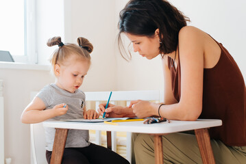 Mom and her beloved daughter drawing together with pencils in children's room, young mother...