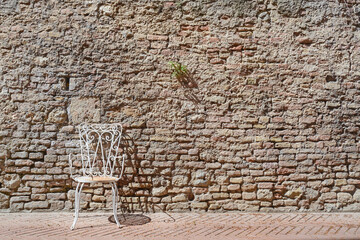 A frontal photo of a white metal chair and a old brick wall in the background in a Italian town 