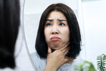worried Asian woman checking her double chin under her lower jaw in front of a mirror, a sign of weight gain or obesity - obrazy, fototapety, plakaty