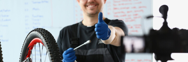 Male blogger holds thumbs up and repairs bicycle remotely. Bike maintenance and live streaming