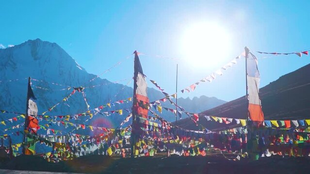 Slow motion shot of prayer flags waving in the wind in front of the snow covered Himalayan mountains and sun on sunny day at Kunzum Pass on the way to Kaza from Manali in Himachal Pradesh, India. 