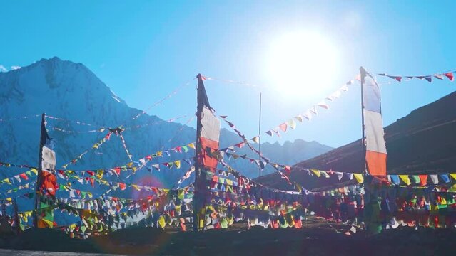 Slow motion shot of prayer flags waving in the wind in front of the snow covered Himalayan mountains and sun on sunny day at Kunzum Pass on the way to Kaza from Manali in Himachal Pradesh, India. 