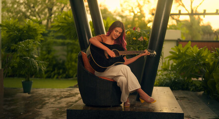 Beautiful Indian woman sitting on chair play acoustic guitar singing alone raining outside home....