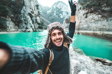 Handsome young man with backpack taking selfie outdoors - Happy hiker standing in front of mountains - Travel blogger enjoying nature view on summer trip - Powered by Adobe