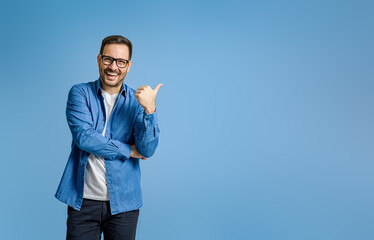 Portrait of cheerful businessman pointing at copy space for advertising against blue background - obrazy, fototapety, plakaty