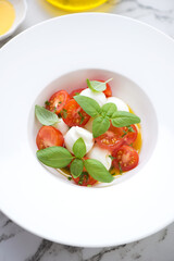 Close-up of caprese salad with mozzarella, red cherry tomatoes, olive oil and fresh green basil served in a white plate, vertical shot