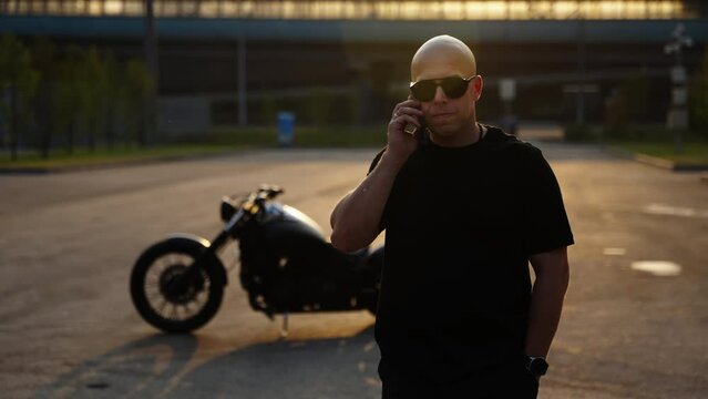 Talking on the phone. A biker against the backdrop of sunset and a black motorcycle walks on the asphalt and holds a phone in his hand.