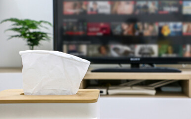 Life at home, close up of box of tissues on white table, television screen on in background out of focus, displaying online streaming platform and their variety of content