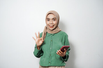 Smiling young Asian Muslim woman dressed in green shirt holding mobile phone and showing okay sign isolated over white background