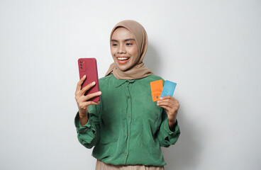Smiling young Asian Muslim woman dressed in green shirt holding mobile phone and credit card, online shopping isolated over white background