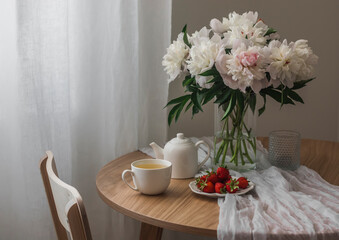Fototapeta na wymiar A bouquet of white peonies in a glass vase, chamomile tea, strawberries on a white round table in a cozy living room