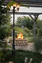 Patio and fire in the fire pit in the evening. A beautiful fire flame, a glowing lantern and a garland against the backdrop of an evening garden.