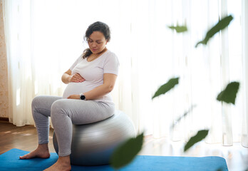 Beautiful pregnant woman, sitting on fitball, holding hands on her belly, exercising in late pregnancy at home, preparing her body for easy childbirth. Sport. Fitness. Active healthy lifestyle concept