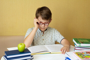 little student boy sitting at table in living room. Redhaired schoolboy learning at home with textbook.