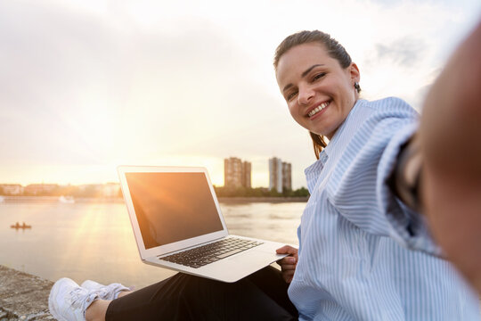 Smiling digital nomad woman working on laptop while sitting on waterfront taking selfie in front of cityscape and sunset.