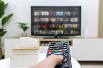 First person view of hand holding remote control, choosing what to watch on streaming platform displayed on television screen in background but out of focus, depicting home life