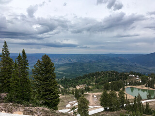 Looking down from peak onto a valley and emerald  lake, pine trees and stormy clouds. View of Wasatch Mountains, Little Cottonwood Canyon, Utah some summer snow left on the ground.
