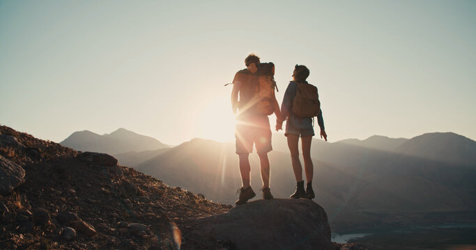 A silhouette of young adventurous couple watching the sunset standing on the top of mountain and holding hands. Traveling together. Happy and freedom concept