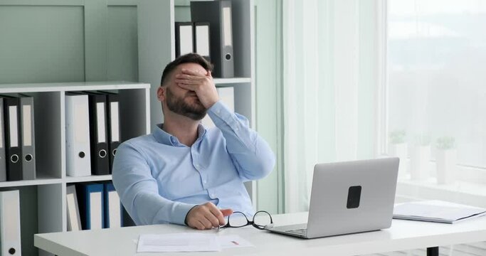 Executive with beard and glasses sitting in office, working on laptop and realizing he made a mistake. He takes off his glasses, rubs his eyes and continues working.