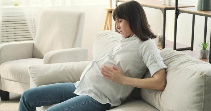 In the comfort of the living room, a weary yet devoted pregnant woman finds solace on the couch. She lovingly caresses her growing belly, emanating warmth and affection, gets up and leaves.