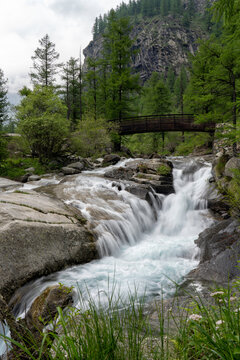 Long exposure of mountain water stream and waterfall in the green forest with a wooden bridge on a cloudy day. Campiglia Soana (Azaria), Gran Paradiso National Park