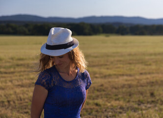 Portrait of young caucasian woman in blue dress with white hat standing on meadow