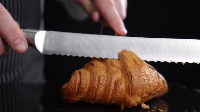 Delicious croissant on black background. Cutting freshly baked french croissant with knife in slow motion. Traditional French cuisine. crumbs falling. puff pastry bakery. Confectionery production