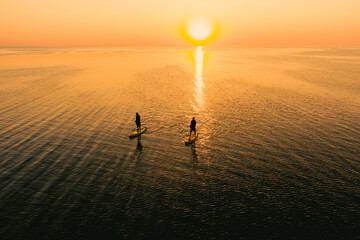 Aerial view of two people on stand up paddle boards on quiet sea at sunset. Warm summer beach vacation holiday.