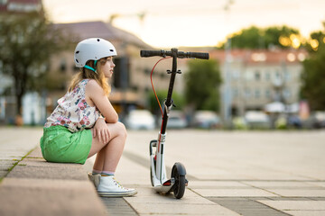 Adorable young girl riding her scooter in a city on sunny summer evening. Pretty preteen child riding a roller. Active leisure and outdoor sports.
