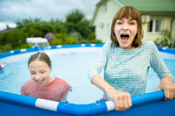 Fototapeta na wymiar Two funny teenage sisters trying to get into cold pool water. Children having fun in outdoor pool. Summer activities for the family with kids.