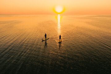 Aerial view of two people on stand up paddle boards on quiet sea at sunset. Warm summer beach...