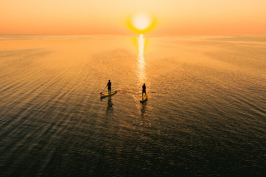 Aerial view of two people on stand up paddle boards on quiet sea at sunset. Warm summer beach vacation holiday.