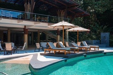 Fototapeta na wymiar Luxurious swimming pool with chaise lounges and sunshades at tropical resort on a bright sunny day, Empty sunbed chairs and parasols arranged around pool