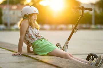 Adorable young girl riding her scooter in a city on sunny summer evening. Pretty preteen child...