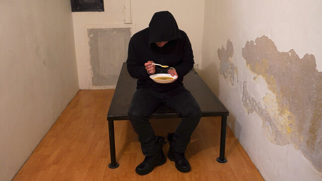 single prison cell, with an iron bed, on which the prisoner sits, eating soup from plastic dishes