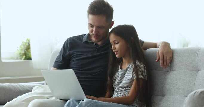 Loving father and 10s pretty daughter sit on sofa in living room use laptop watch family movies, spend weekend together on internet, discussing online purchase. Young generation use modern technology