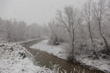 Winter landscape with snow coming down and a river in the middle of trees covered by snow - 611780446